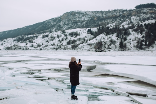 Une fille dans une veste d'hiver noire, prenant des photos du paysage d'hiver de la rivière gelée avec des glaces.