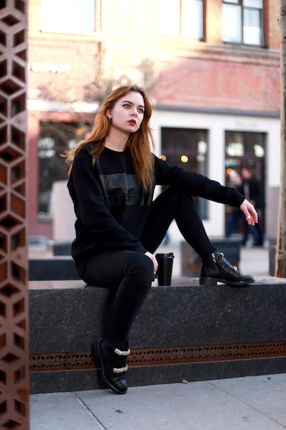 Fille dans des vêtements élégants noirs s'assoit à l'extérieur avec du café. Style de la ville. Modèle aux cheveux roux.