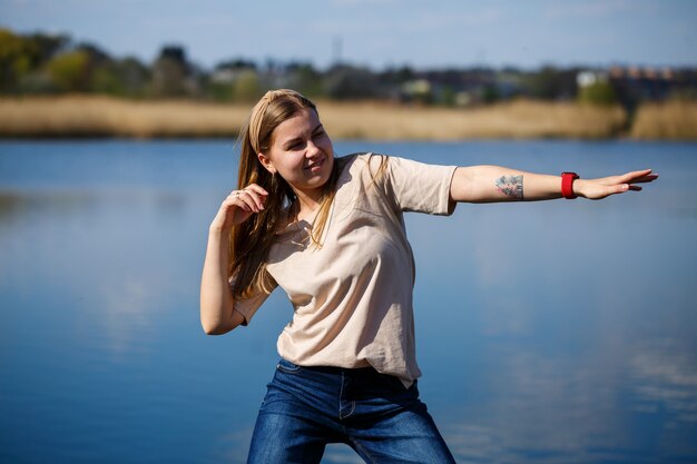 Fille dansant près du lac, temps ensoleillé. Une jeune femme se réjouit de la vie, danse et chante. Elle est de bonne humeur et le sourire aux lèvres.