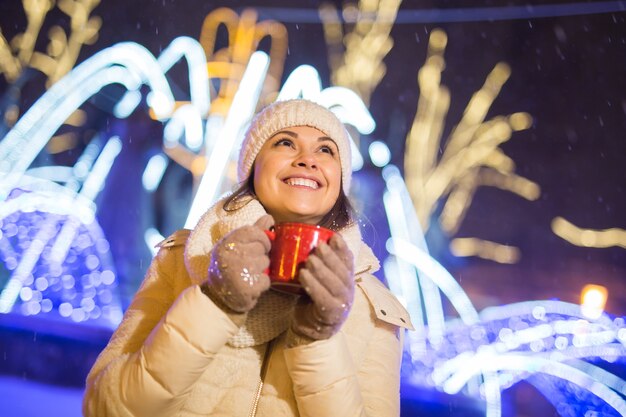 Fille debout sur fond de ville de noël hiver neige neige dérive, se tient chapeau de veste chaude, tient une tasse de thé avec des boissons au thé chaud.