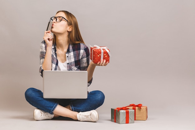 Une fille décontractée excitée tenant un ordinateur portable et une carte de crédit alors qu'il était assis sur un sol avec pile de coffrets cadeaux