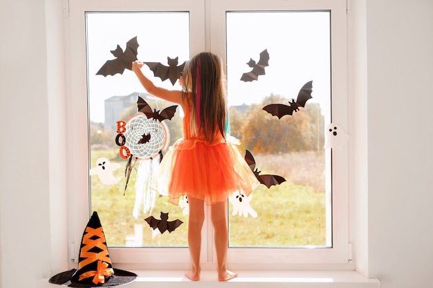 La fille décore la fenêtre de la pièce avec des chauves-souris et des fantômes d'Halloween debout sur le rebord de la fenêtre