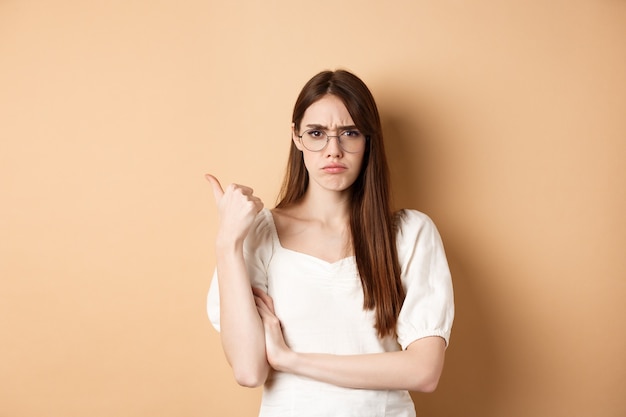 Fille déçue dans des verres fronçant les sourcils, pointant de côté le mauvais produit, désapprouve et n'aime pas quelque chose, debout sur beige.