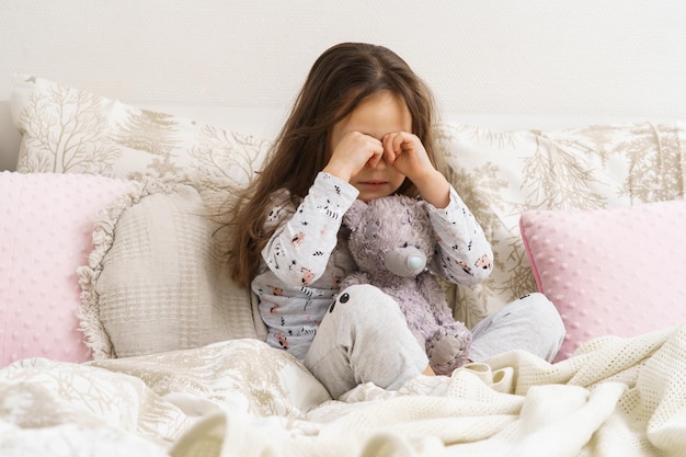 Fille déçue déprimée se frottant les yeux s'asseoir avec un ours en peluche dans un lit blanc et doux le matin Enfants orphelins