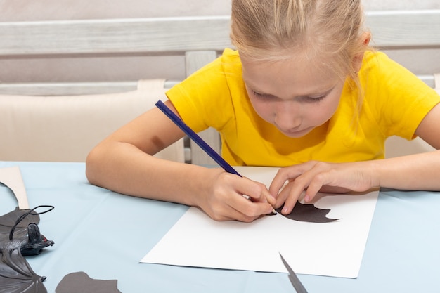 Une fille dessine et découpe une décoration d'Halloween à la maison dans du papier noir. Décorations artisanales de bricolage. L'enfant fait des chauves-souris noires en papier, origami