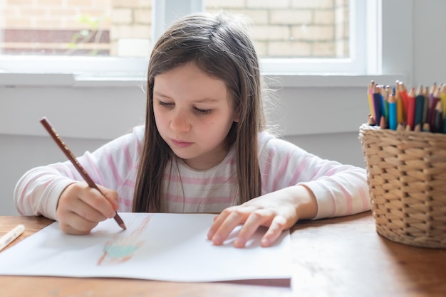 Une fille dessine sur du papier alors qu'elle est assise sur la table à la maison.