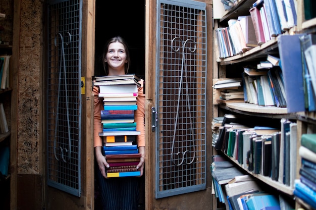 Fille détient une grande pile de livres et porte beaucoup de littérature dans la bibliothèque