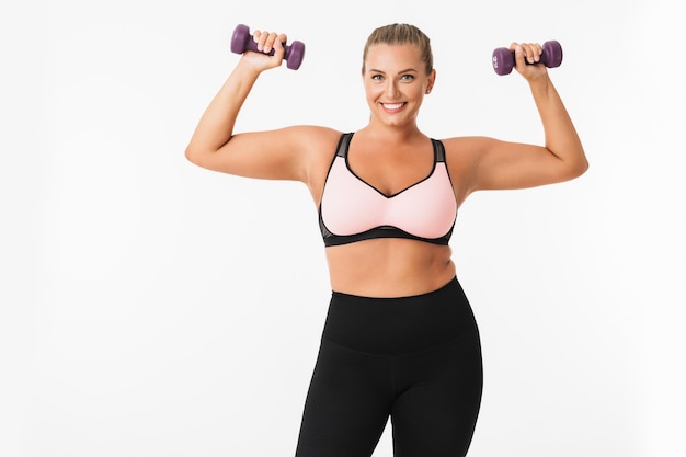 Fille dodue souriante en haut sportif et leggings tenant des haltères dans les mains tout en regardant joyeusement à huis clos sur fond blanc