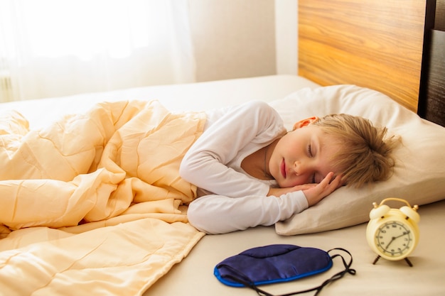 Une fille dort dans son lit, tôt le matin, le soleil brille dans la fenêtre et à côté du réveil. Un sommeil sain.