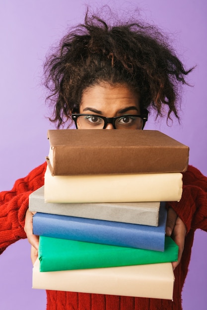 Fille drôle afro-américaine en uniforme scolaire tenant un tas de livres, isolé