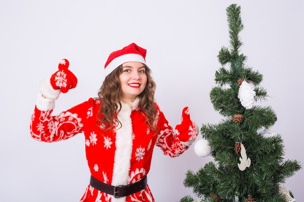 Fille drôle en costume de père Noël près de sapin de Noël montrant le geste de pouce en l'air.