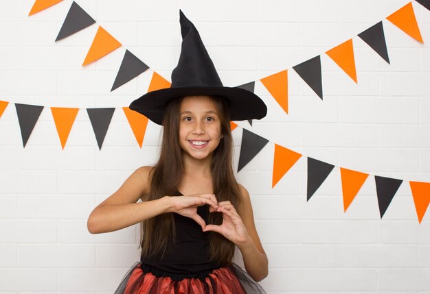 La fille drôle drôle dans un chapeau et un costume de sorcière pour Halloween montre le coeur avec ses doigts