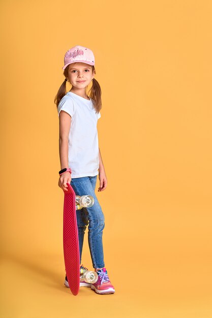 Fille drôle élégante portant un t-shirt blanc, un jean bleu et des baskets, tenant une planche à roulettes sur un mur jaune