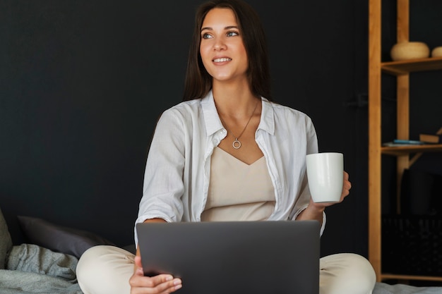 Fille du millénaire est assise sur le lit en chemise blanche, tenant une tasse de café à la main.