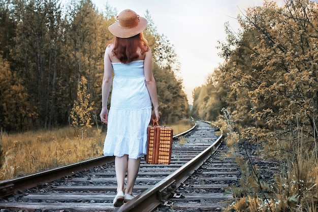 fille du parc d'automne dans une robe d'été blanche et une valise en osier marchant sur des rails