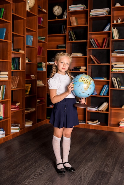 Fille de l'école intelligente en uniforme se tient avec un globe dans la bibliothèque de l'école