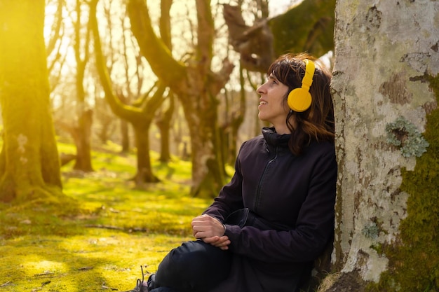 Une fille écoutant de la musique avec un casque jaune dans la forêt au coucher du soleil assis sur un arbre