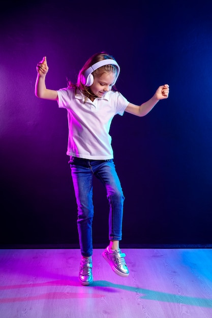 Fille écoutant de la musique dans les écouteurs sur un mur coloré sombre. Danseuse. Heureuse petite fille dansant sur la musique. Enfant mignon appréciant la musique de danse heureuse.