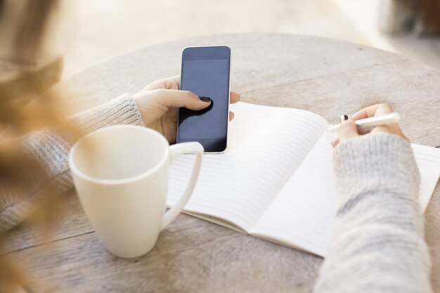 La fille écrit dans un cahier avec le téléphone portable et la tasse de café