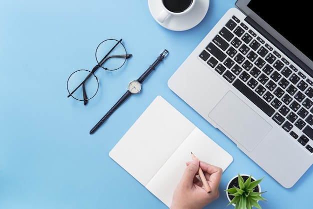 Une fille écrit sur un livre blanc ouvert ou une comptabilité sur un bureau bleu clair minimal et propre avec un ordinateur portable et des accessoires, espace de copie, vue de dessus à plat, maquette