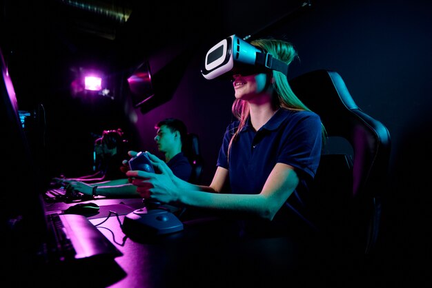 Fille eenage contemporaine dans le casque vr à l'aide du joystick alors qu'il était assis devant un écran d'ordinateur et jouant à un nouveau jeu vidéo en club