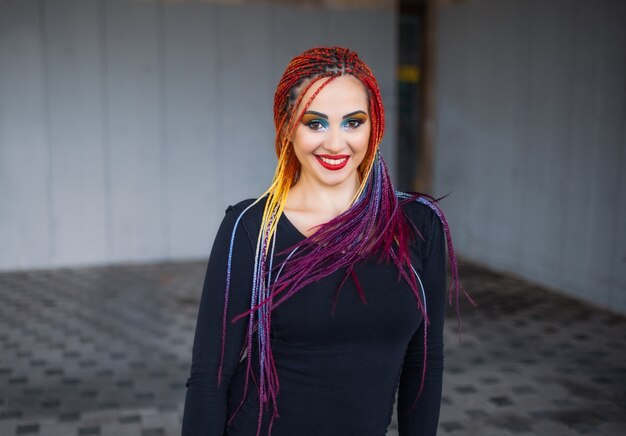 Fille élégante inhabituelle dans une robe simple noire avec un maquillage lumineux et des tresses multicolores posant