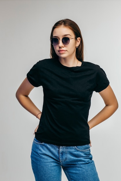 Fille élégante à lunettes portant un t-shirt noir posant sur un mur gris