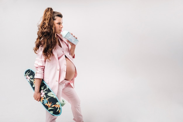 Une fille enceinte en costume rose avec une planche à roulettes dans ses mains boit du jus sur fond gris.