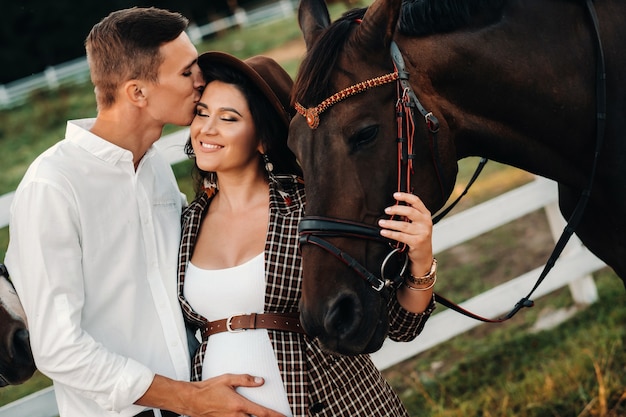 Une fille enceinte dans un chapeau et un homme en vêtements blancs se tiennent à côté de chevaux près d'une clôture blanche. Femme enceinte élégante avec un homme avec des chevaux. Couple marié.