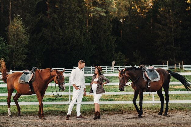 Une fille enceinte dans un chapeau et un homme en vêtements blancs se tiennent à côté de chevaux près d'une clôture blanche.