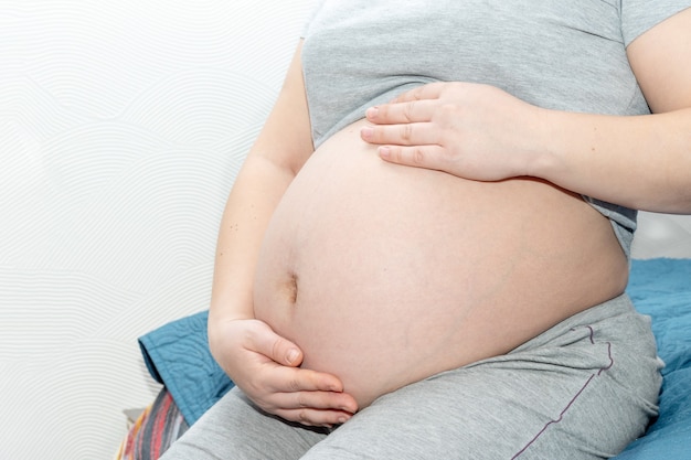 Une fille enceinte est assise sur le lit à la maison et tient ses mains sur son ventre. Concept de grossesse et de famille.