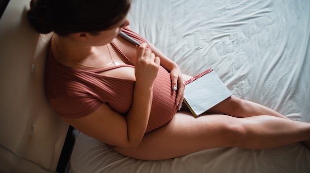 Une fille enceinte est assise sur un lit et repasse son ventre avec un cahier sur ses genoux. Liste à la maternité. En attendant le bébé. Place pour le texte. Espace de copie
