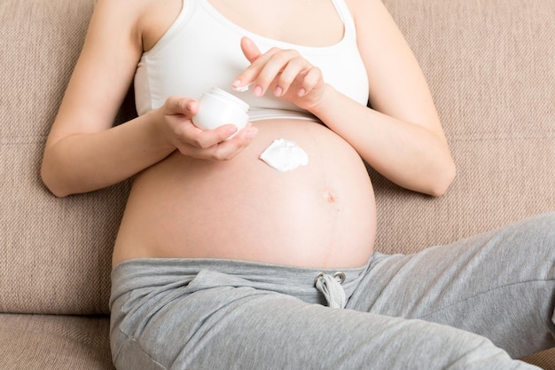 Une fille enceinte est assise à la maison sur le lit et enduit le sourire d'une crème anti-vergetures sur le ventre Préparation à la grossesse et concept d'attente