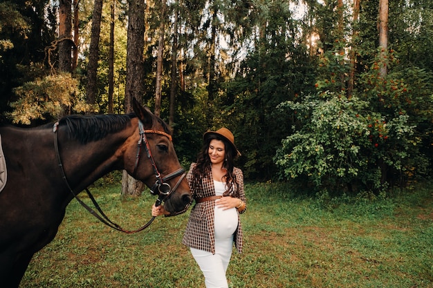 Fille enceinte avec un gros ventre dans un chapeau à côté de chevaux dans la forêt dans la nature