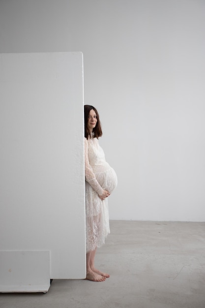 une fille enceinte avec un gros ventre regarde derrière un mur blanc