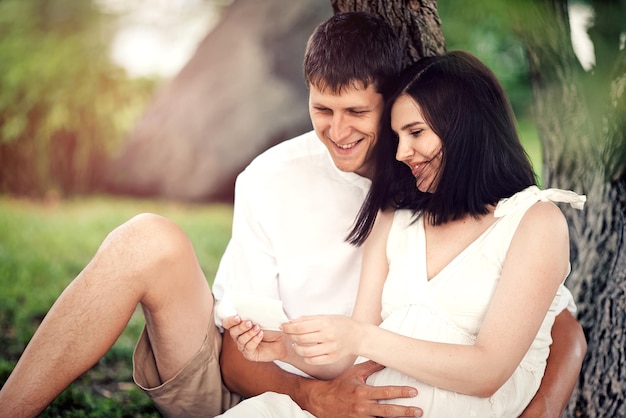 Une fille enceinte et un homme sont assis sous un arbre dans le parc et regardent la photo échographique de l'enfant à naître