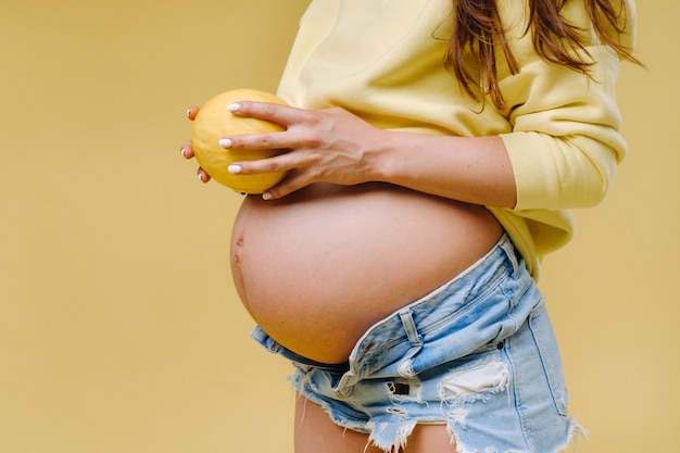 Une fille enceinte avec une veste jaune se tient avec un melon dans ses mains sur un fond jaune isolé