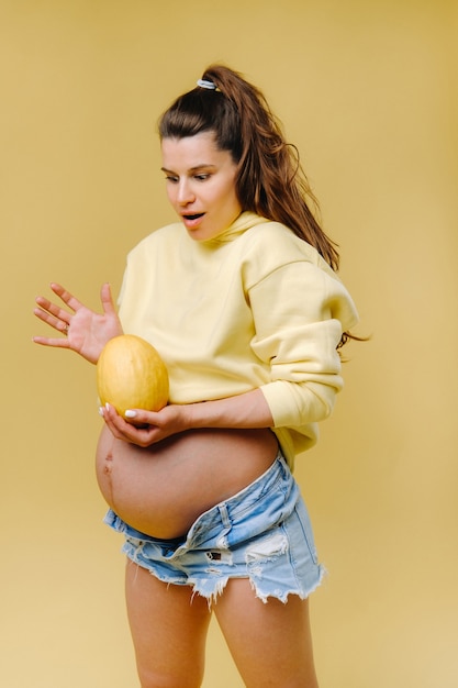 Une fille enceinte avec une veste jaune se tient avec un melon dans ses mains sur un fond jaune isolé