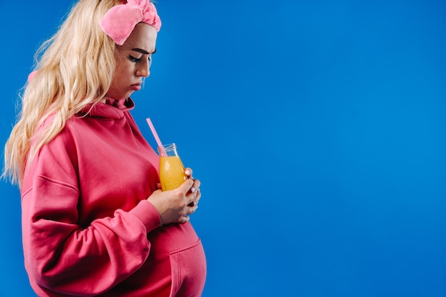 Fille enceinte en vêtements roses avec une bouteille de jus sur fond bleu.