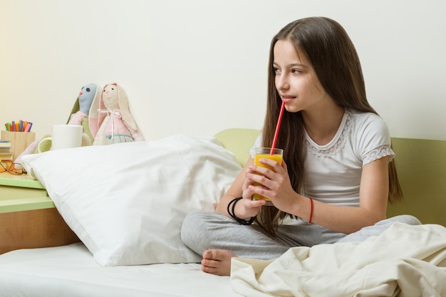 Une fille endormie est assise dans son lit à la maison et boit du jus d'orange