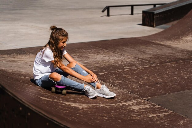 Fille enfant active après la chute d'un penny board blessée assise et ressentant de la douleur sur la rampe de sport sur l'aire de jeux du skate park