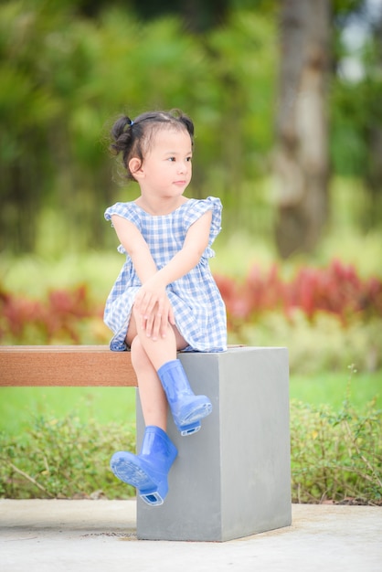 Fille d'enfant asiatique heureuse dans le fond d'arbre de jardin de parc, Bel enfant s'amusant à jouer dehors avec un sourire heureux enfants jouant à l'extérieur portrait de petite fille porter des bottes colorées banc assis