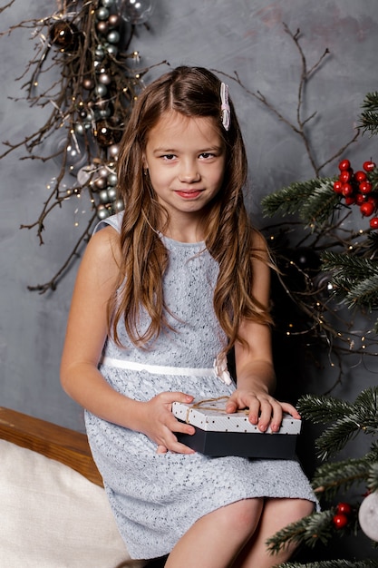 Fille enfant assise près de l'arbre de Noël la veille de Noël, tenant une boîte-cadeau