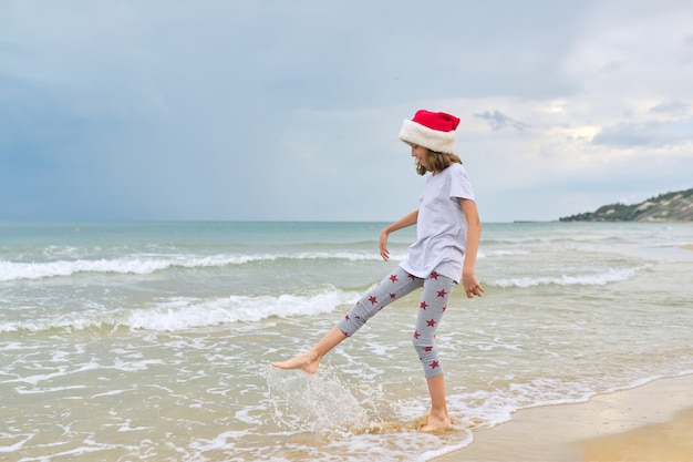 Fille enfant en bonnet de Noel s'amusant sur une plage de l'océan