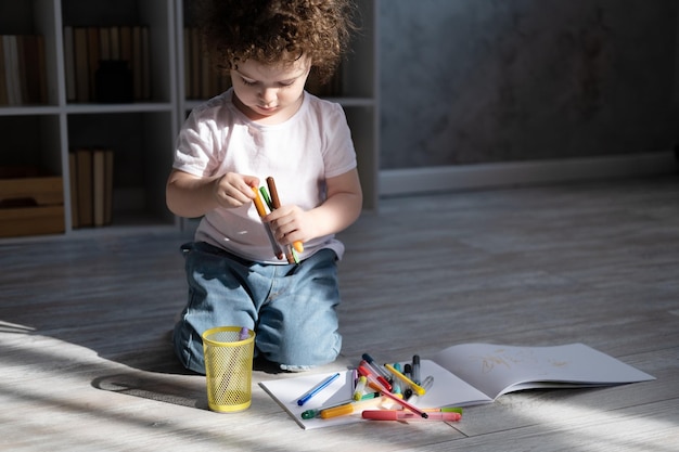 Fille enfant bouclée dessinant avec des marqueurs de couleur assis sur le sol dans le salon à la maison