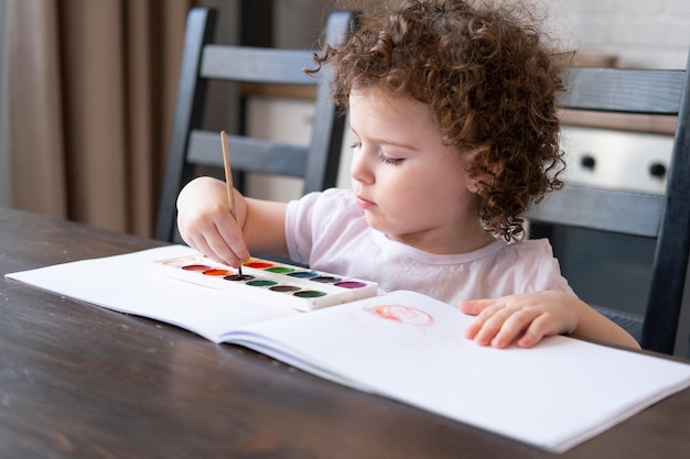 Fille enfant bouclée peignant avec des peintures à l'aquarelle à la maison à la table de la cuisine