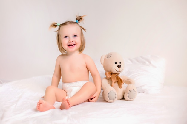 Fille enfant en couches est assis avec ours en peluche sur le lit à la maison