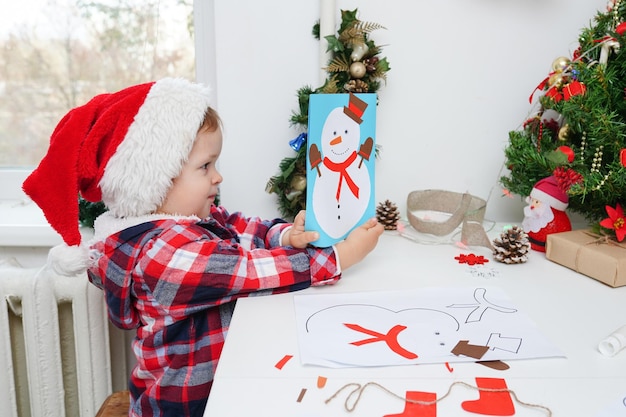 Fille d'enfant dans le chapeau de Santa faisant la carte de voeux de Noël avec le bonhomme de neige