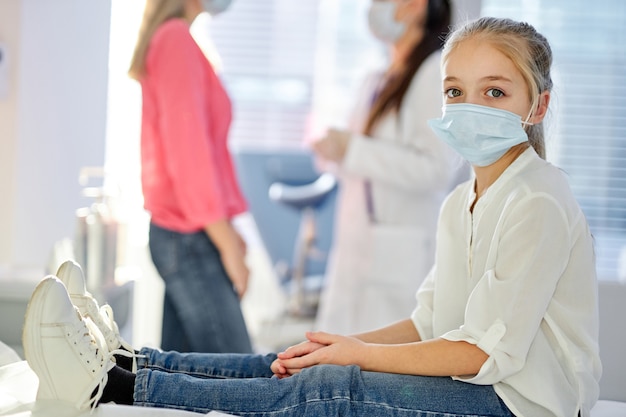 Fille enfant dans un masque médical regardant la caméra, assis sur le lit à l'hôpital ou à la clinique pendant que sa mère parent parle avec l'infirmière en arrière-plan