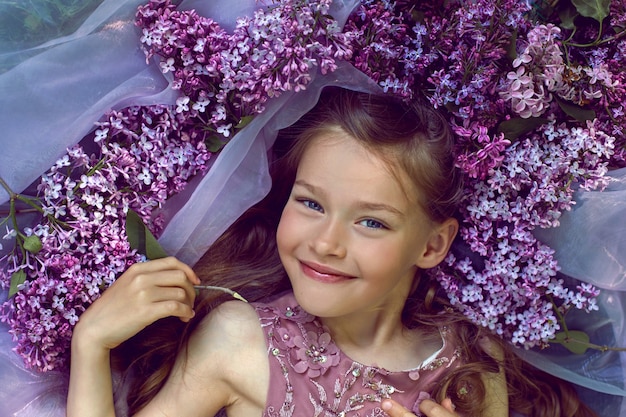 Fille enfant dans une robe à fleurs violette se trouve sur le sol parmi les lilas sur un voile au printemps